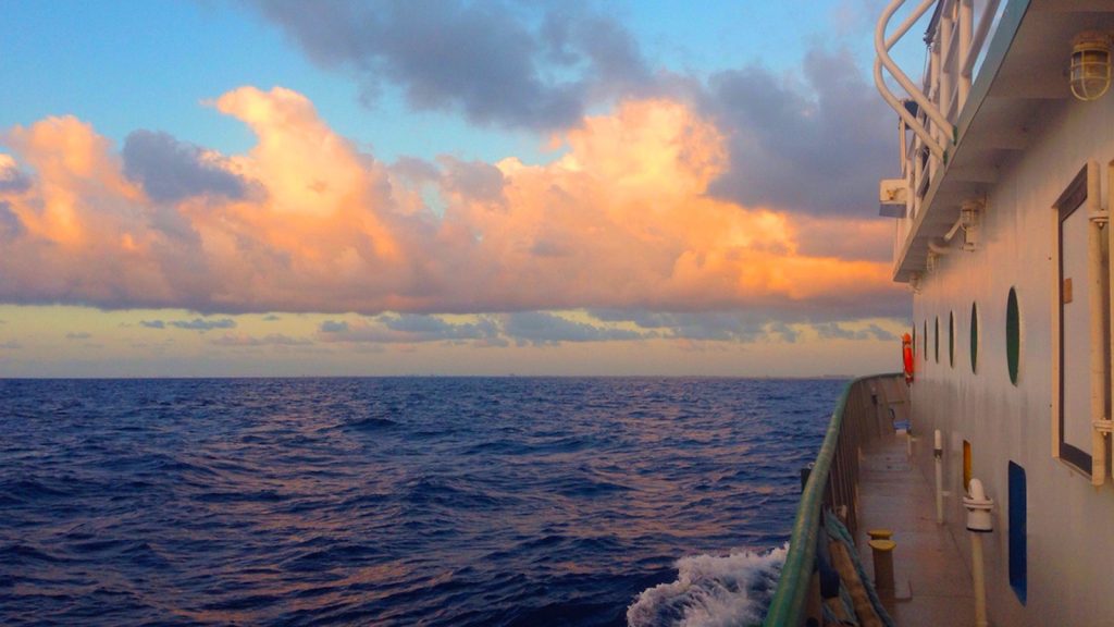 Light orange and pinkish hue highlight the passing clouds beneath a beautiful blue sky and vast open ocean with small movements and the single splash of whitewater against the hull of the Walton Smith that takes up only the right edge of the photo, the image taken from the deck as the sun rises during a research cruise, the green gunnel and white wall of the Walton Smith cabin aligned with portholes in view