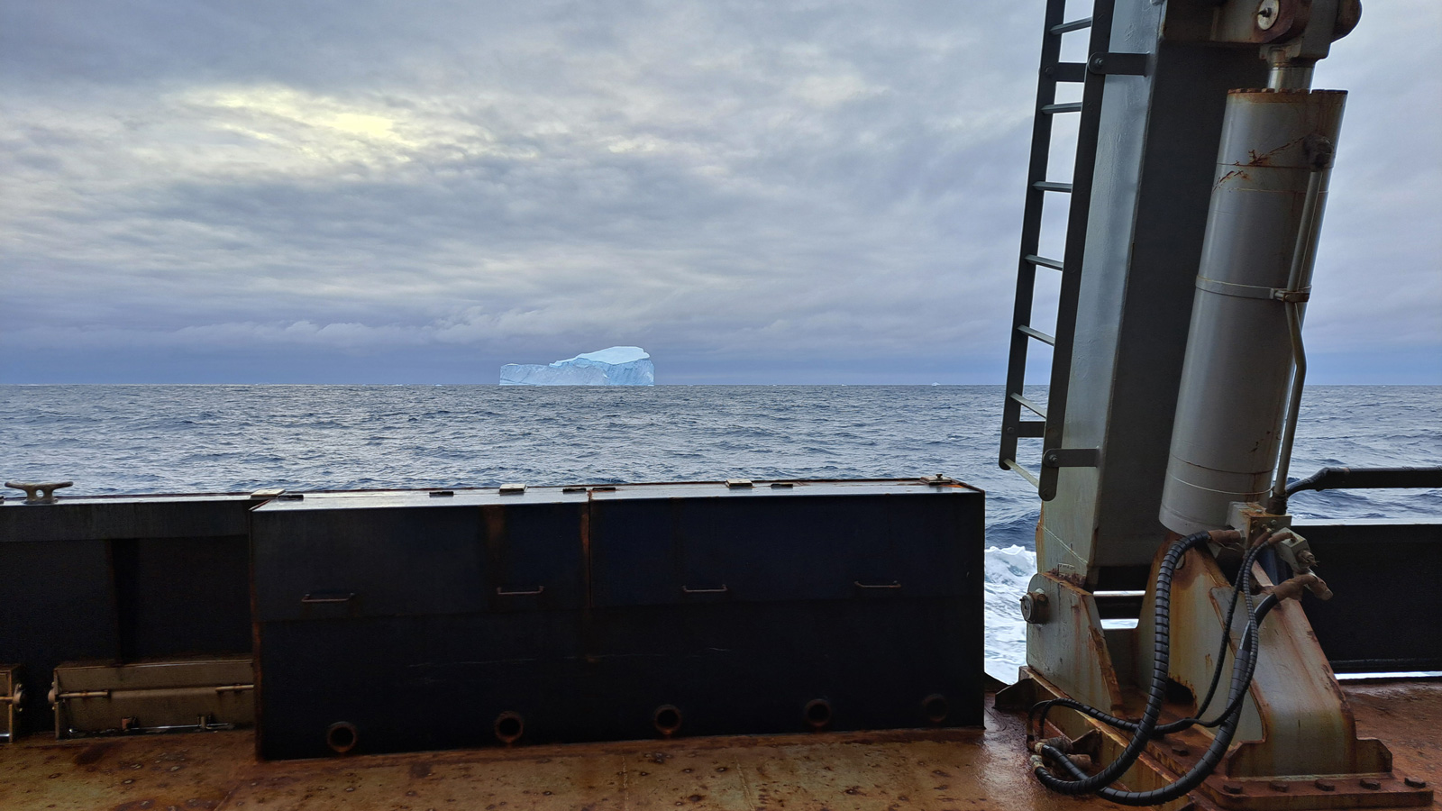 A blue and white iceberg at the center of the photo in the open blue ocean taken from the side of the R/V Marcus Langseth with the rusty gunnel and white crane at the bottom and right of the photo
