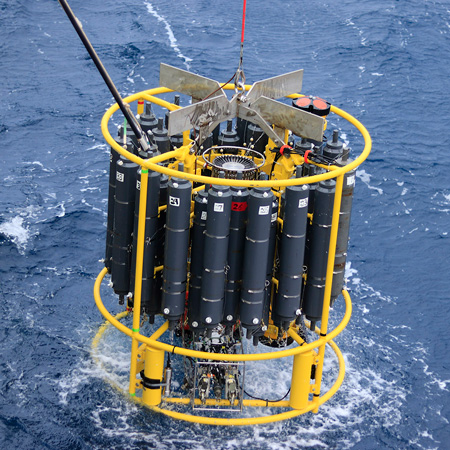 A yellow rimmed CTD with black Niskin bottles in a rosette dives into the blue ocean creating white water on the I08S GO-SHIP Cruise