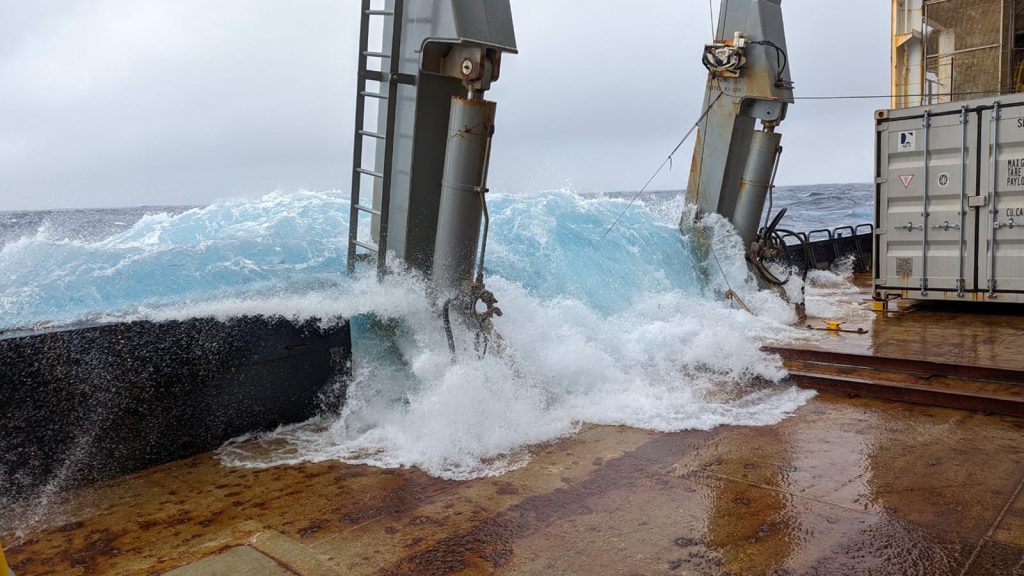 A light blue massive wave pours over the rusty huge gunnel of the R/V Marcus Langseth with grey clouds while storm hits the A13.5 GO-SHIP CRUISE