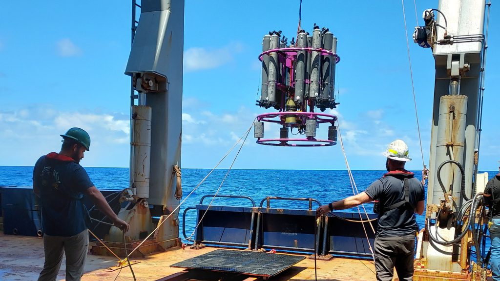 Ian Smith (left) and Jay Hooper (right) both in dark blue te shirts, khaki pants , a white hard hat standing on the rusty starboard side of the R/V Marcus Langseth holding a set of ropes as the massive CTD Rosette with gray long niskin bottles positioned in a circular pattern is suspended in the airover blue ocean on the side of the boat between the two white pilings that compose the CTD's deployment A13.5 
