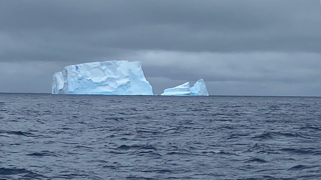 A massive iceberg with a piece separating off to the right in the deep blue ocean beneath the darkening sky and the gray clouds