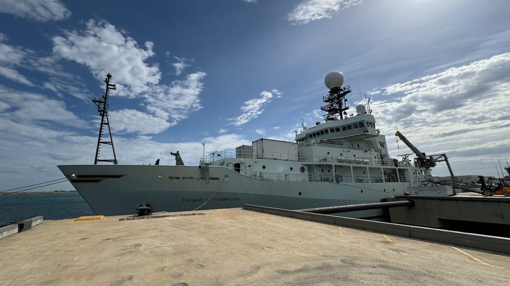 THE R/V Thomas Thompson (silver and gray) at dock in Fremantle with towers and three decks beneath a blue sky and white clouds as the sun beams down on the ship on the I08S GO-SHIP CRUISE