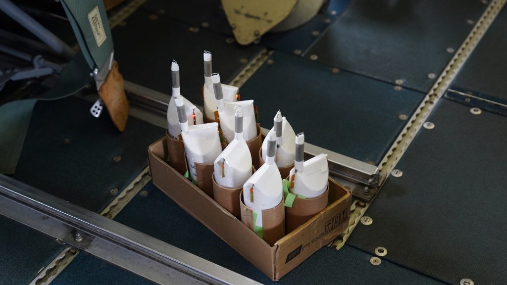 Image shows the white Skyfora Streamsondes sitting in rows inside a cardboard box. They are sitting on the floor of the P3 aircraft, and are waiting to be released from the chute of the aircraft.