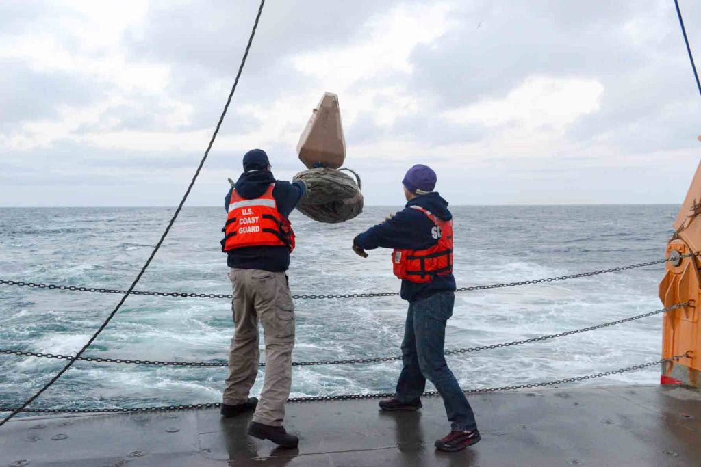 Two people at the stern of a research vessel throw a global ocean drifter into the waves below.